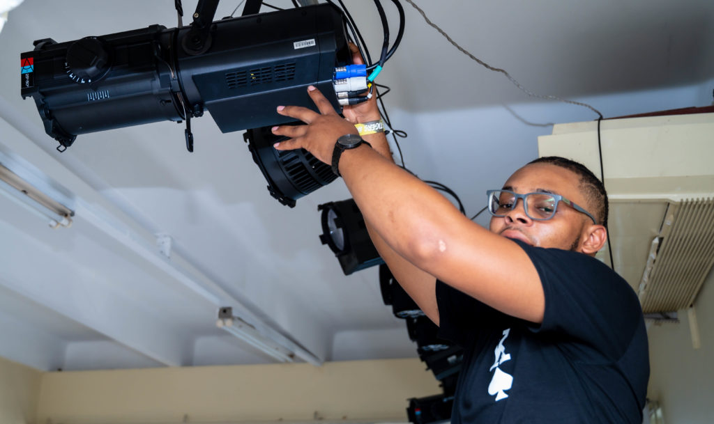 Lighting design student smiles for the camera while adjusting lighting equipment donated by the Advance Program
